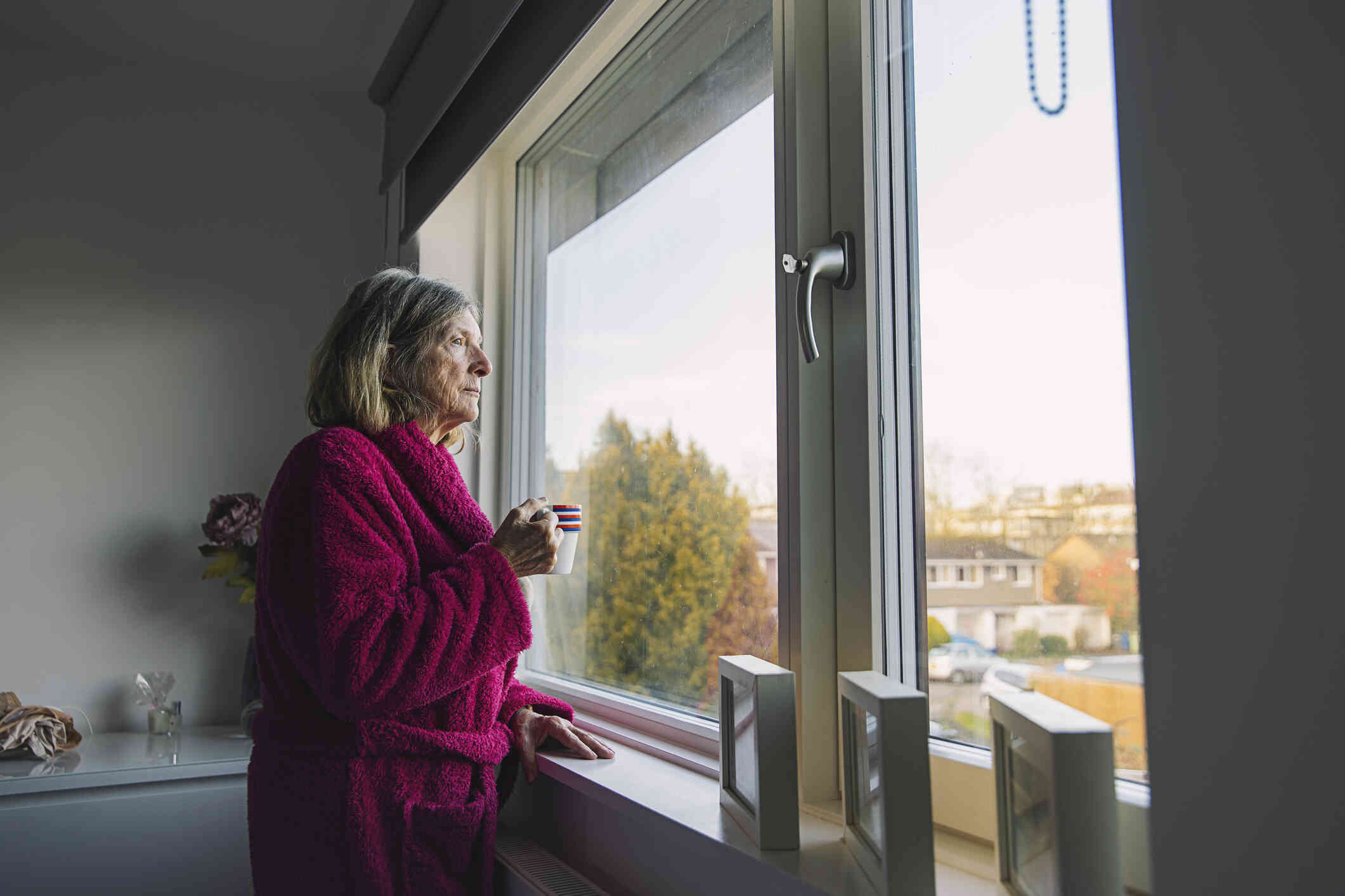 An elderly woman in a red houserobe stands in her kitchen and gazesd sadly out of the kitchen window while holding a coffee mug in her hand.